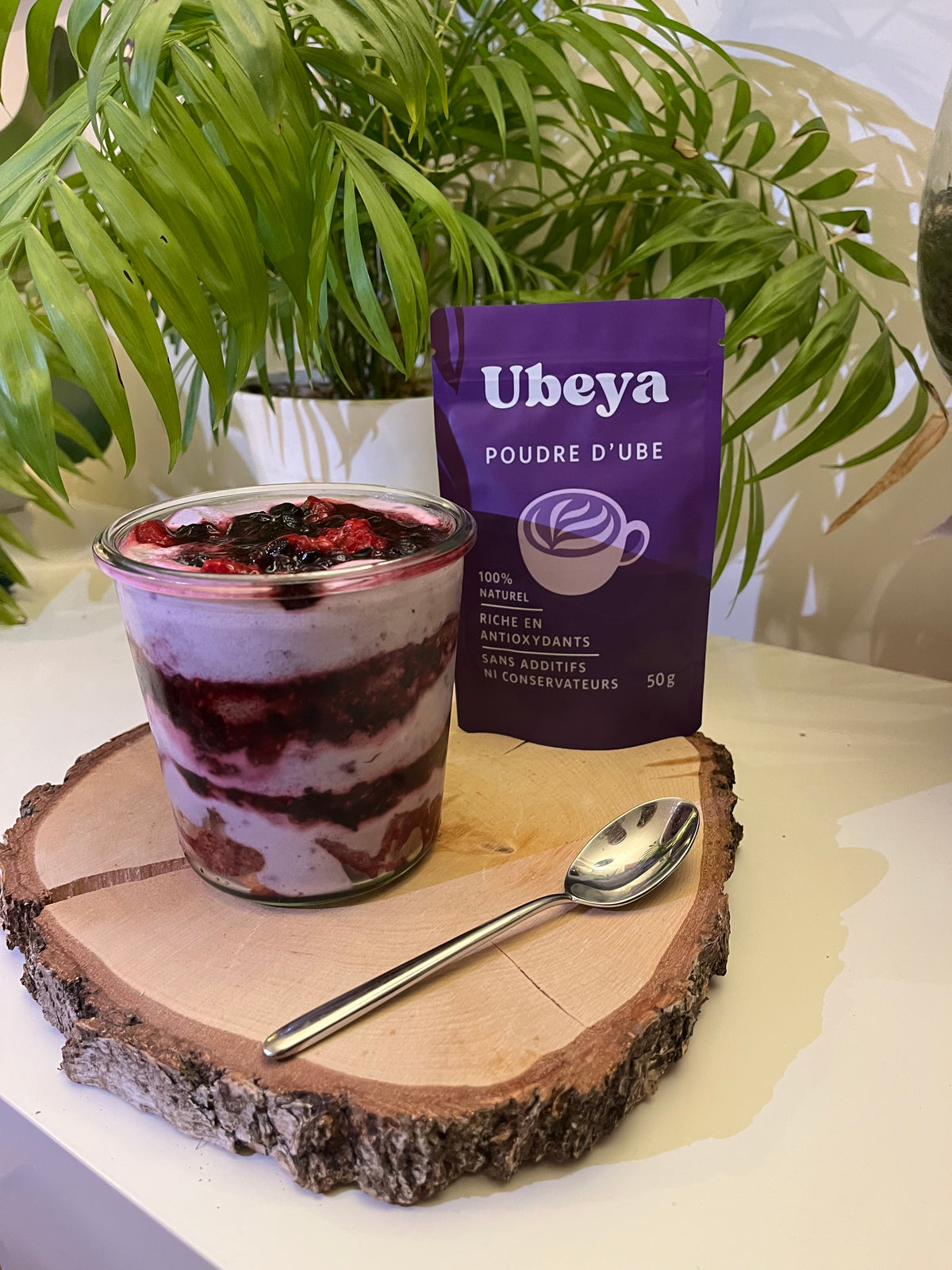 Sachet Úbeya debout à côté d'un croissant fourré à la crème d'ube et d'une tasse de latte violet, fond clair, lumière naturelle latérale.